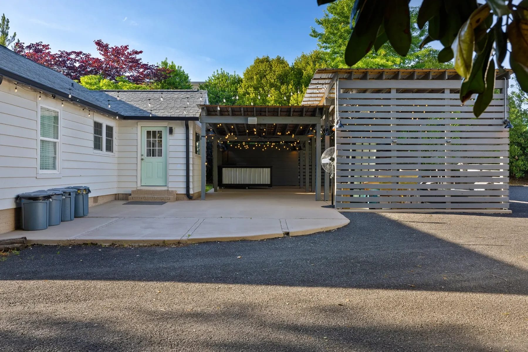 A white house with a light green door opens onto a covered patio area with string lights and a slatted wooden pergola beside a sectioned-off structure. Four trash bins line the wall on a paved driveway. Trees fill the background. Huntersville event venue