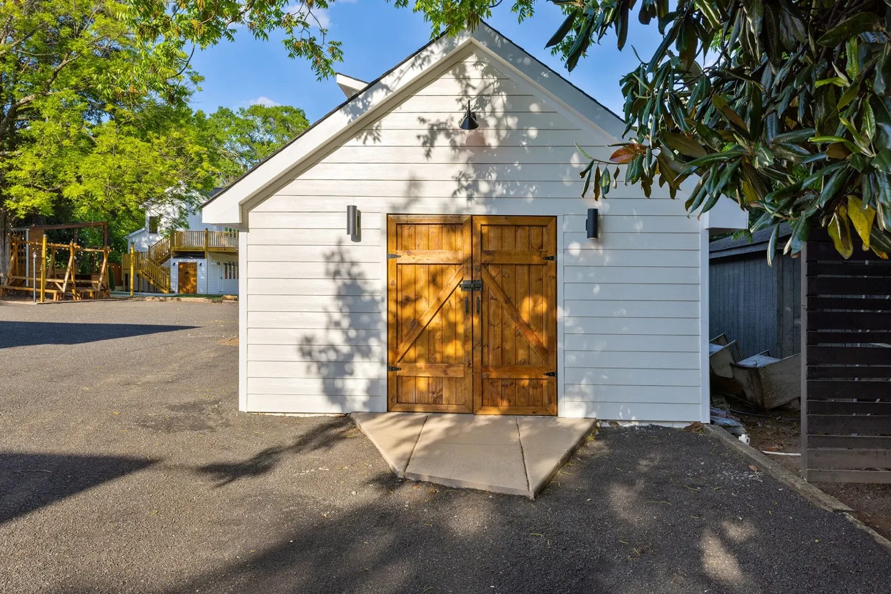 A small white building with a peaked roof and double wooden doors sits on an asphalt surface. Trees cast shadows on the building, and greenery surrounds the area. Huntersville event venue