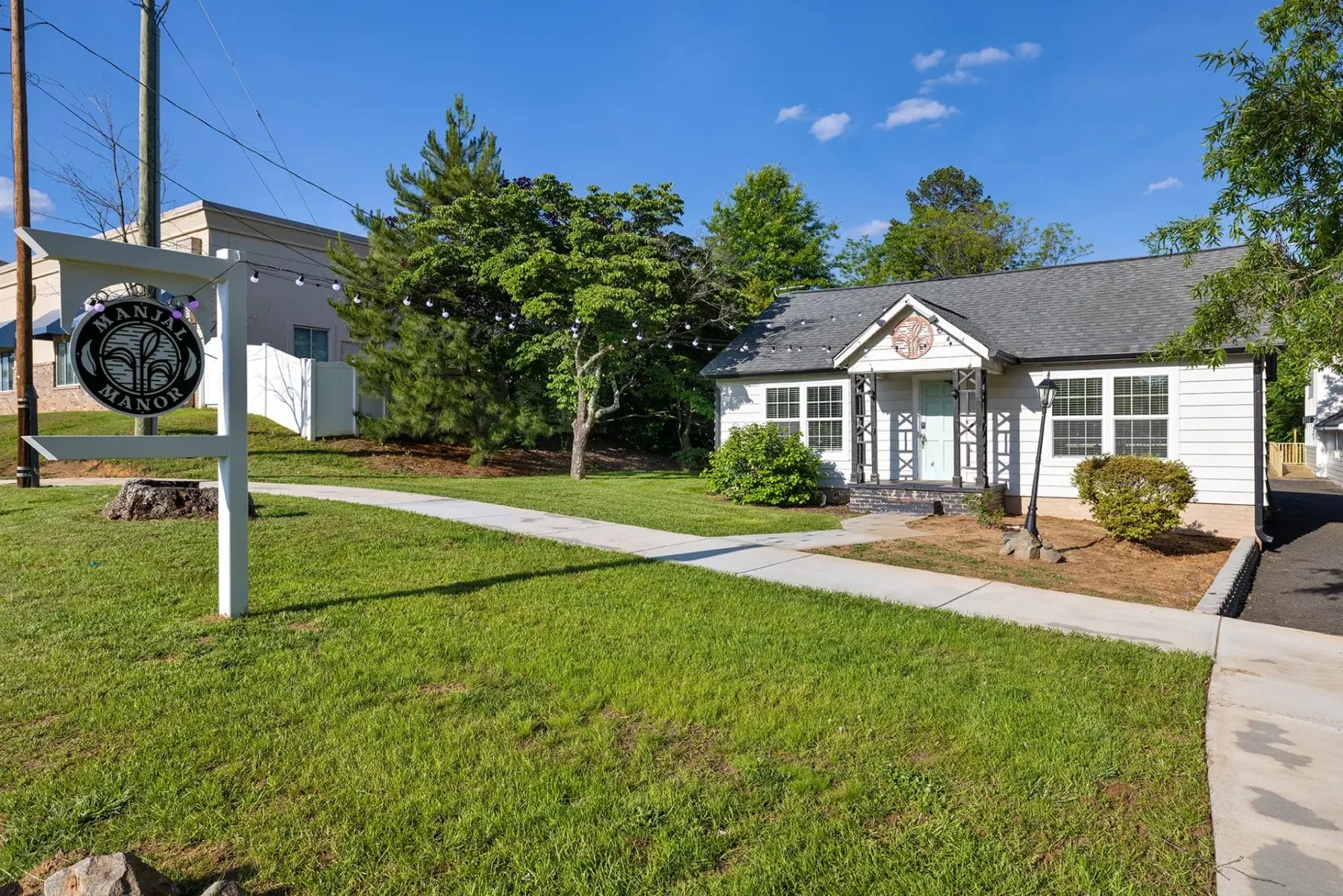 First Look: Exciting New Transformations at Manjal Manor 81 A small white building with black trim and a gray roof sits by a sidewalk, surrounded by green grass, trees, and bushes. A white sign with a black circular logo stands on the lawn near the street. Huntersville event venue