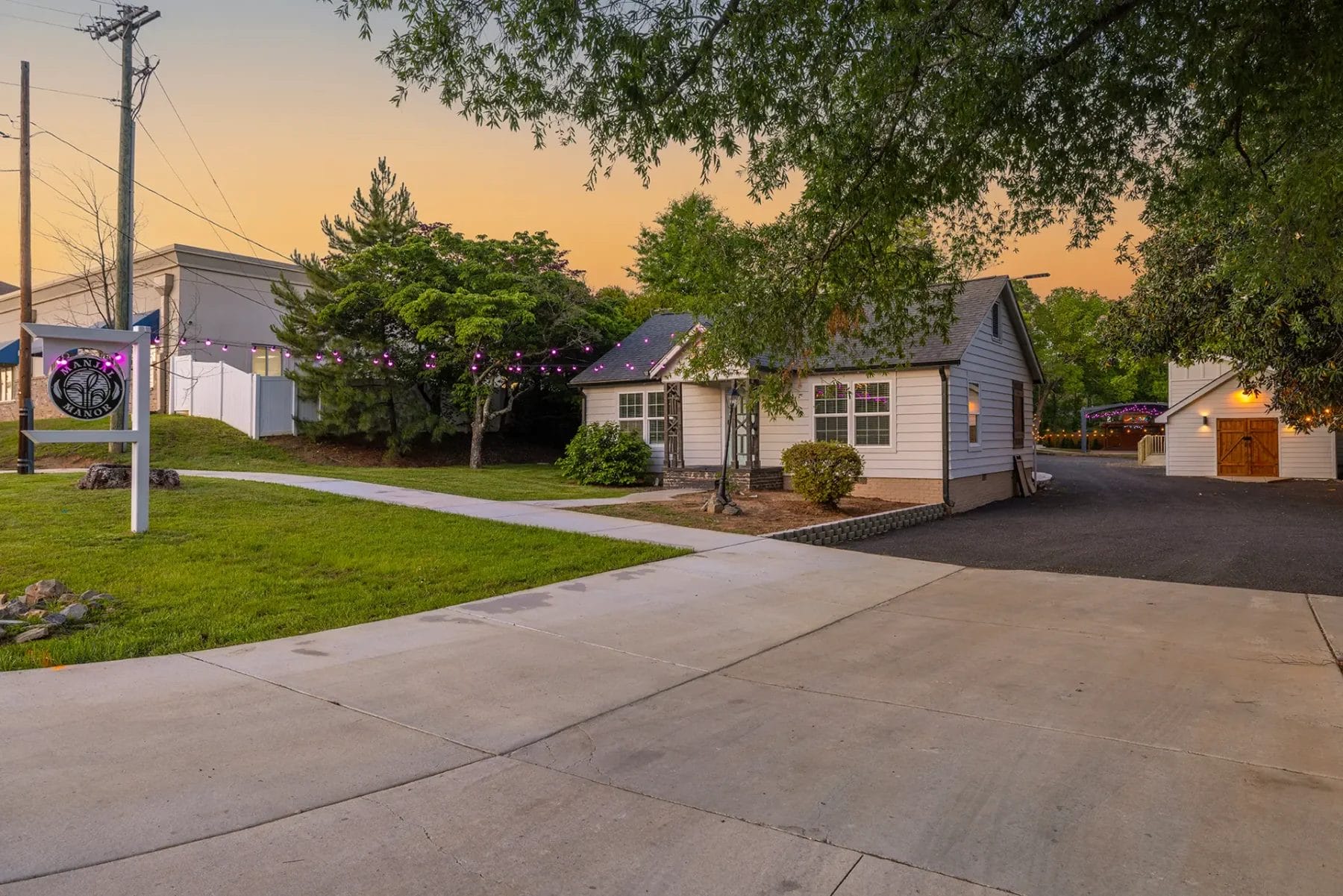 A small house with string lights on the roof sits by a wide driveway at sunset, surrounded by green grass, shrubs, and trees. A signpost is visible on the left side of the image. Huntersville event venue