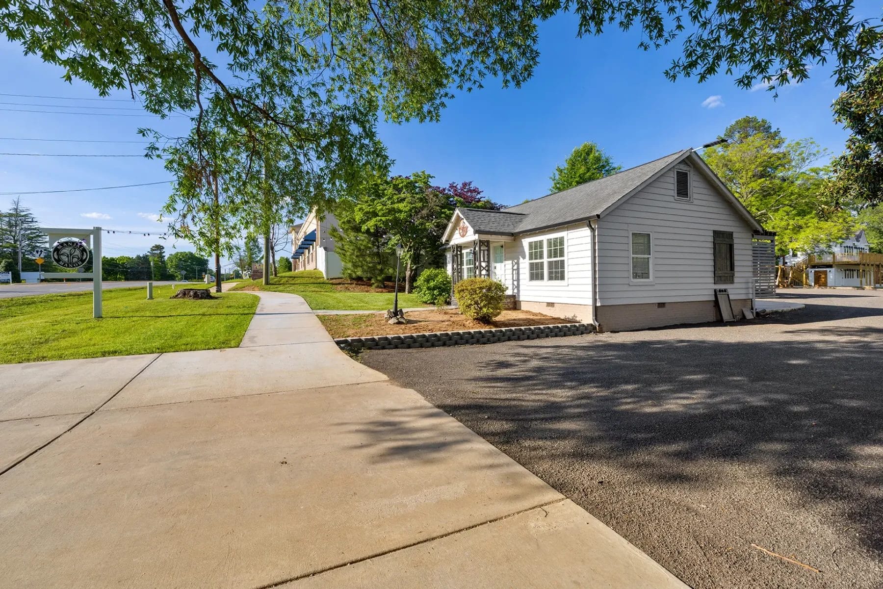 A small white house with gray roof sits beside a sidewalk, surrounded by trees and greenery under a clear blue sky on a sunny day. The driveway and a signpost are visible in the foreground. Huntersville event venue
