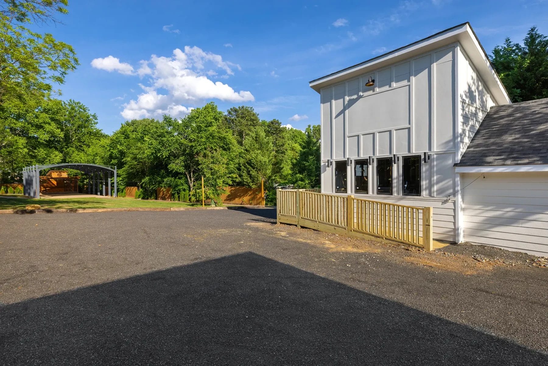 A modern white building with large windows and a wooden deck sits beside an asphalt driveway, surrounded by trees under a blue sky with scattered clouds. A metal carport is visible in the background on the left. Huntersville event venue