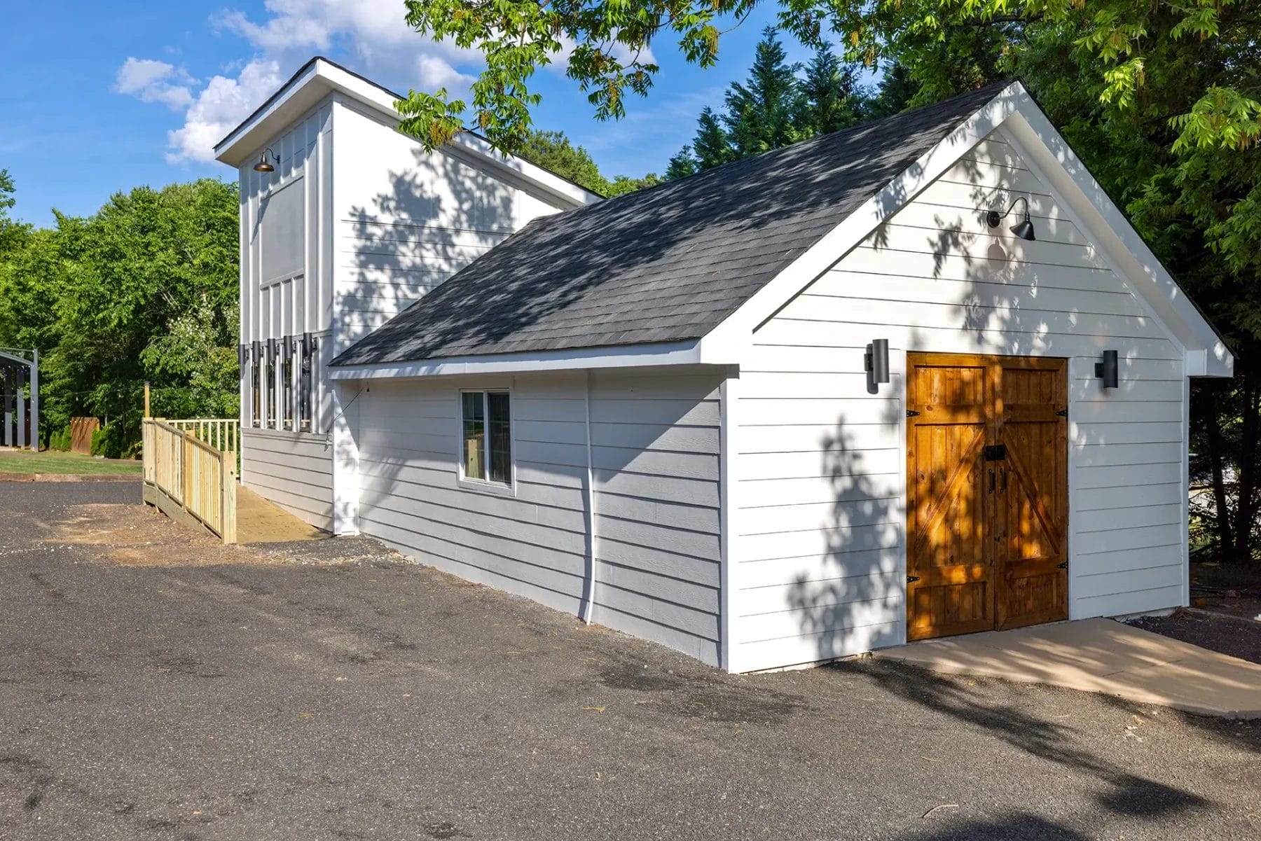 A modern white barn-style building with a sloped shingle roof, wooden double doors, and a ramp on the side, surrounded by trees and set against a clear blue sky. Huntersville event venue