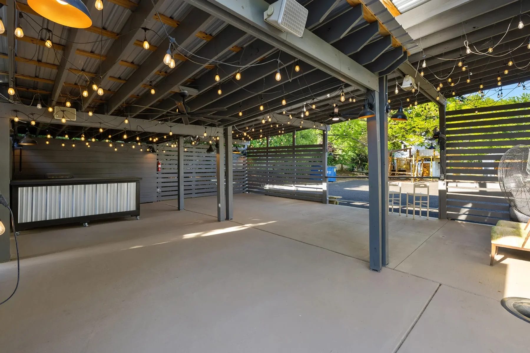 First Look: Exciting New Transformations at Manjal Manor 83 A covered outdoor patio with string lights hanging from the ceiling, gray metal beams, slatted privacy panels, and a concrete floor. Natural light enters from the open sides, and trees are visible outside. Huntersville event venue