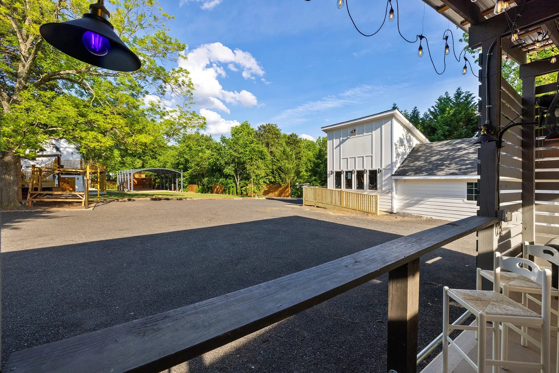View from a shaded porch with string lights, overlooking a paved courtyard, white building, playground, and trees under a blue sky with scattered clouds. Huntersville event venue
