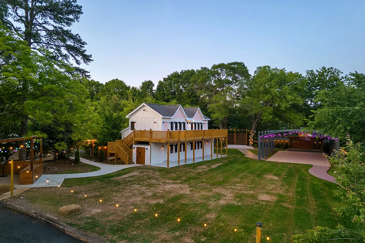 First Look: Exciting New Transformations at Manjal Manor 23 A large white two-story house with a wraparound wooden deck sits among trees. Nearby, a covered outdoor area and pathways are decorated with string lights. The grassy yard has scattered landscape lights. Huntersville event venue