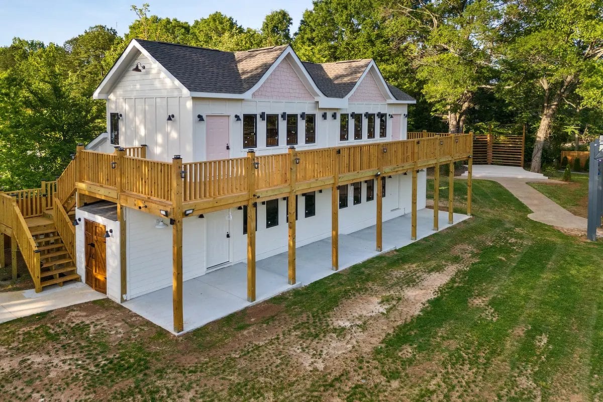 A two-story white house with a large wooden deck and staircase, elevated on tall wooden supports, surrounded by green trees and a grassy yard. Huntersville event venue
