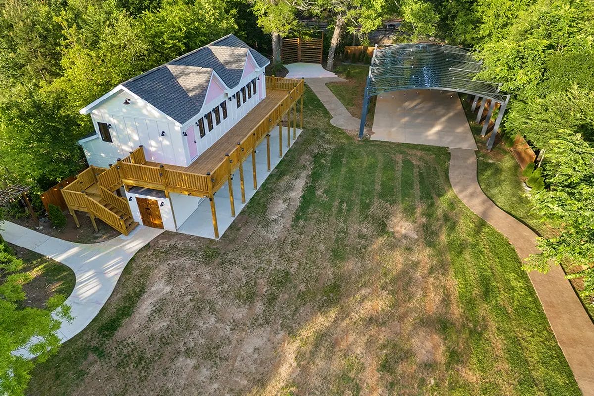 Aerial view of a white building with a wooden deck and stairs, surrounded by trees, a lawn with patchy grass, curved walkways, and a covered open-air structure nearby. Huntersville event venue