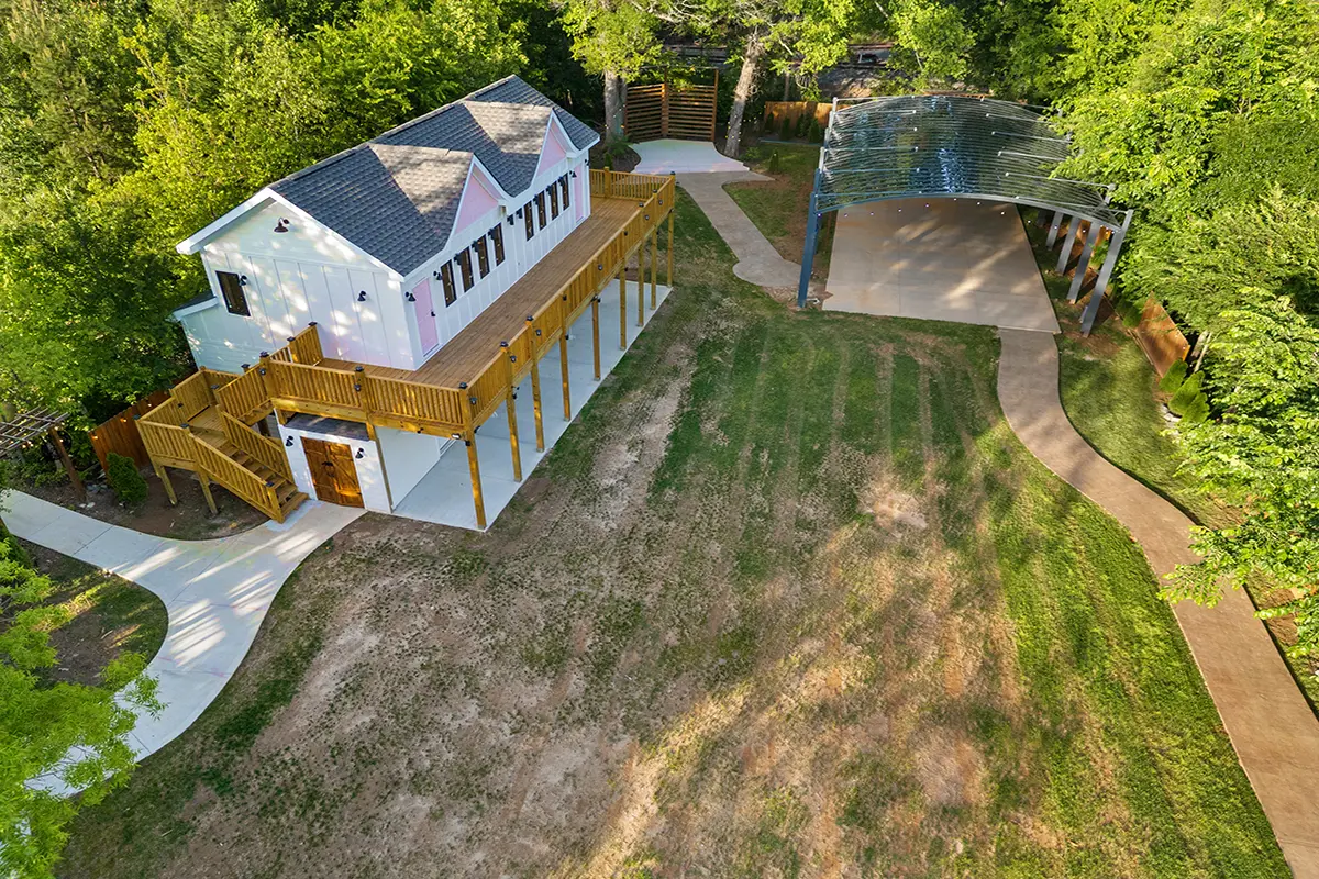 First Look: Exciting New Transformations at Manjal Manor 48 Aerial view of a white building with a wooden deck and stairs, surrounded by trees, a lawn with patchy grass, curved walkways, and a covered open-air structure nearby. Huntersville event venue
