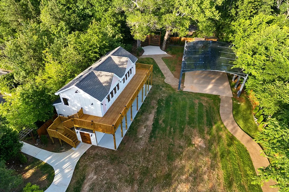 Aerial view of a white two-story house with a large wooden deck, surrounded by green trees, a lawn with some bare patches, winding concrete paths, and a metal-roofed pavilion nearby. Huntersville event venue
