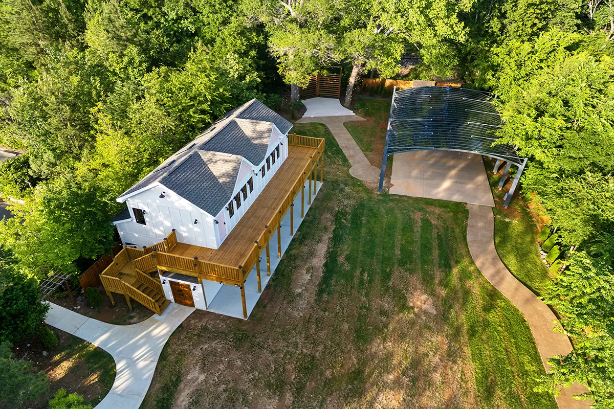 First Look: Exciting New Transformations at Manjal Manor 32 Aerial view of a white two-story house with a large wooden deck, surrounded by green trees, a lawn with some bare patches, winding concrete paths, and a metal-roofed pavilion nearby. Huntersville event venue