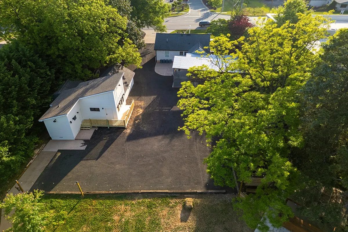 Aerial view of a small white house with a deck and a gray roof, surrounded by trees and greenery. The house sits next to a large paved area and another building, with a road visible in the background. Huntersville event venue
