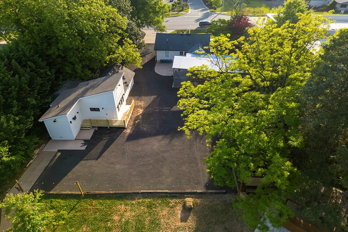 First Look: Exciting New Transformations at Manjal Manor 37 Aerial view of a small white house with a deck and a gray roof, surrounded by trees and greenery. The house sits next to a large paved area and another building, with a road visible in the background. Huntersville event venue