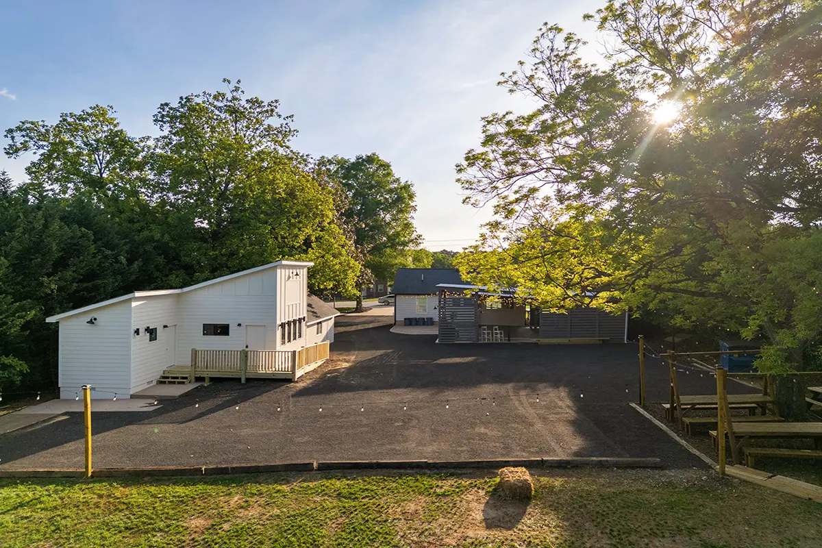 First Look: Exciting New Transformations at Manjal Manor 36 A small modern white building with a deck sits in a paved lot, surrounded by trees in bright sunlight. Other small structures and wooden fencing are visible in the background. Huntersville event venue