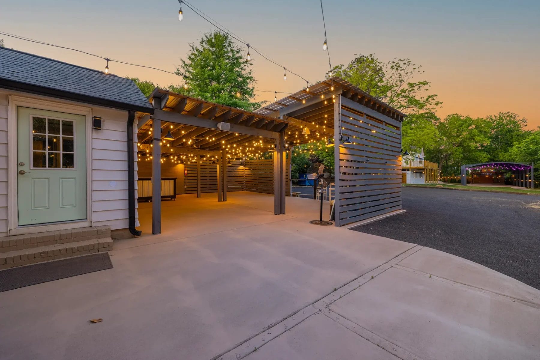 Covered patio with string lights at sunset, attached to a white house with a green door. The area features a modern pergola and paved surface, surrounded by trees and a driveway. Huntersville event venue