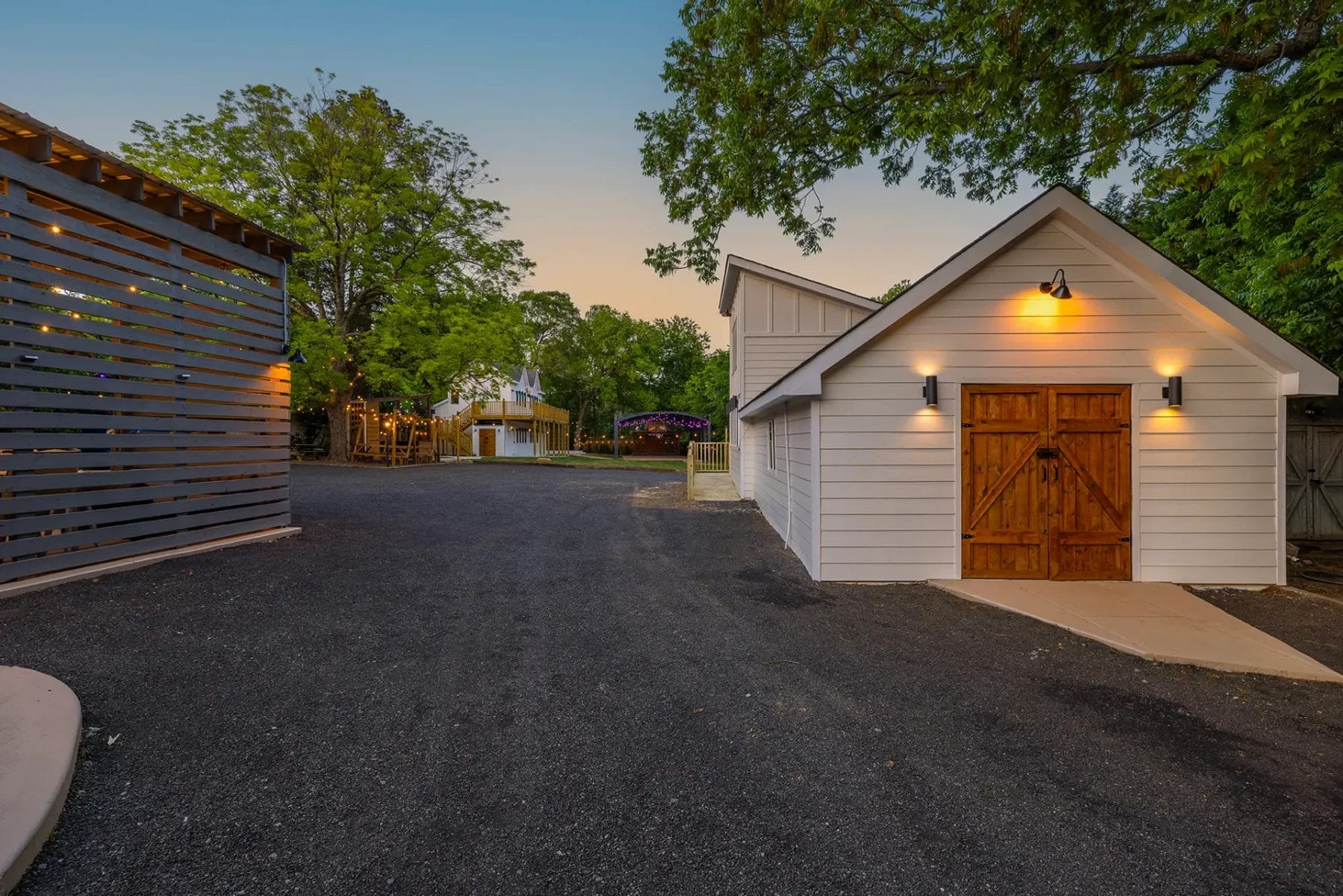 A white barn-style building with wooden doors and warm outdoor lights, set on a paved driveway with trees and other structures visible in the background at sunset. Huntersville event venue