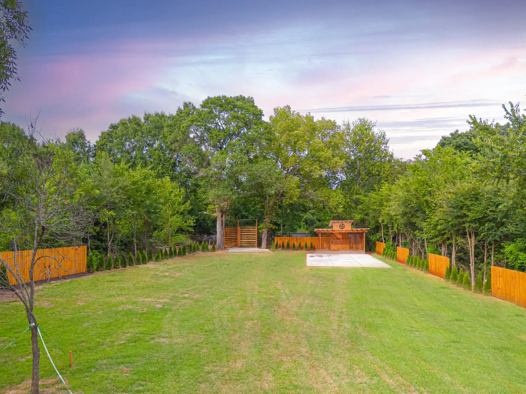 Nestled in Huntersville, this spacious backyard offers a well-maintained lawn surrounded by trees and a wooden fence, perfect as an enchanting wedding venue. A small wooden structure and paved area on the right enhance its charm against the backdrop of a colorful sky. Huntersville event venue