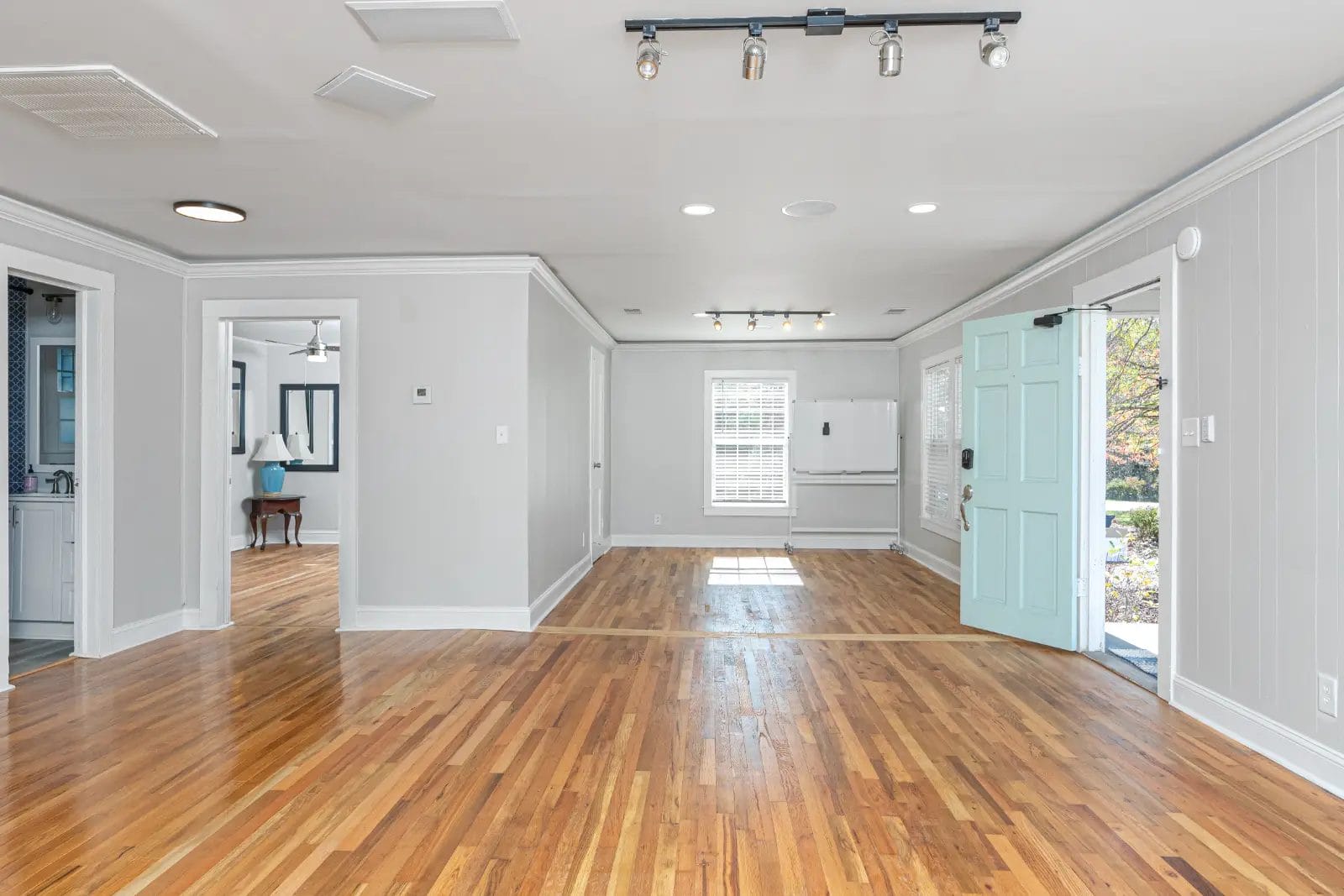 A bright, empty room with hardwood floors and light gray walls serves as a versatile meeting space for rent. An open turquoise door sits on the right, with sunlight streaming through a large window. Recessed lighting and a track light adorn the ceiling. A small mirrored room is visible on the left. Huntersville event venue
