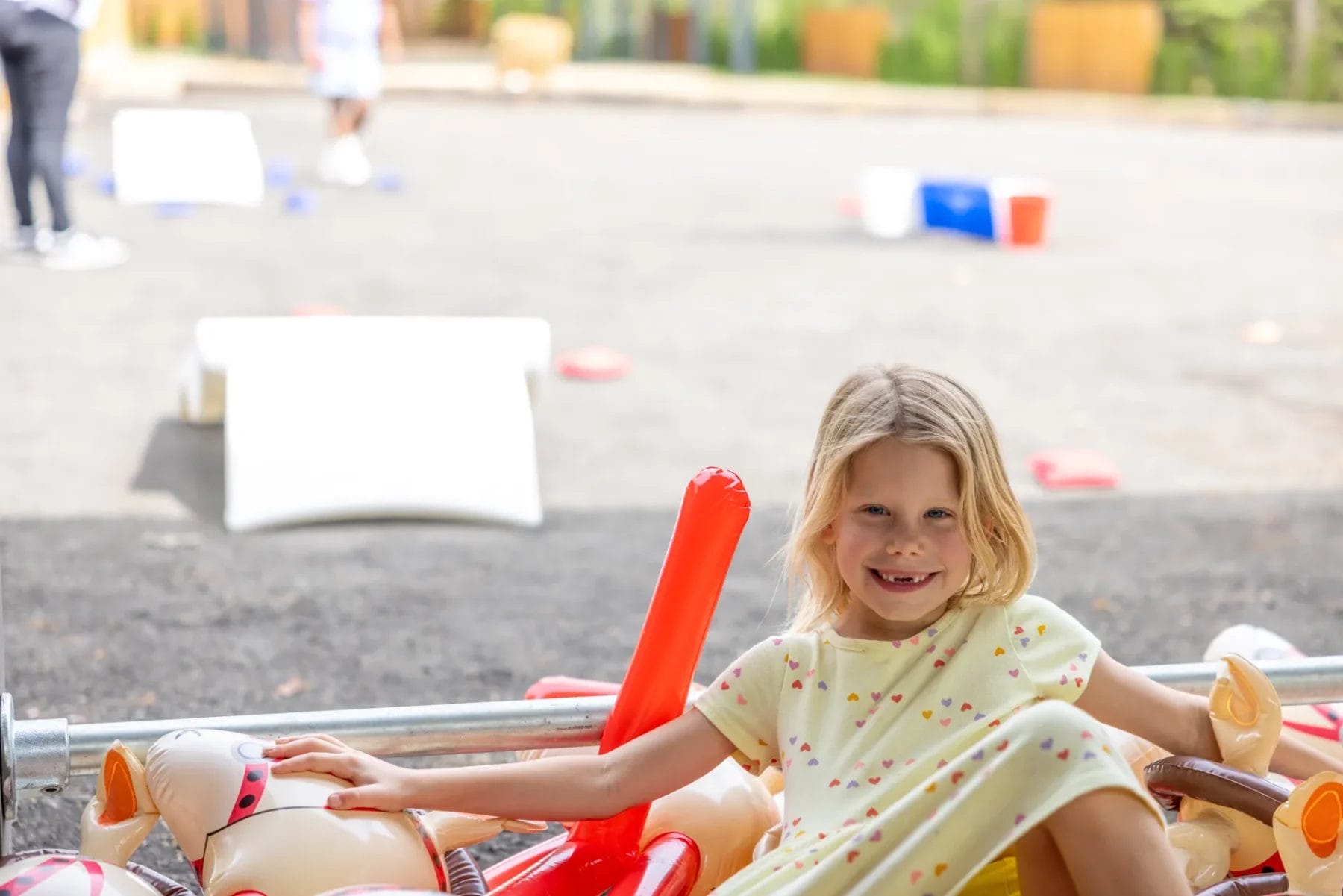 A young girl with light brown hair smiles while sitting on inflated toys outdoors. She wears a light yellow dress with small colorful patterns. The background shows a playground area with ramps and a blurry figure. Huntersville event venue