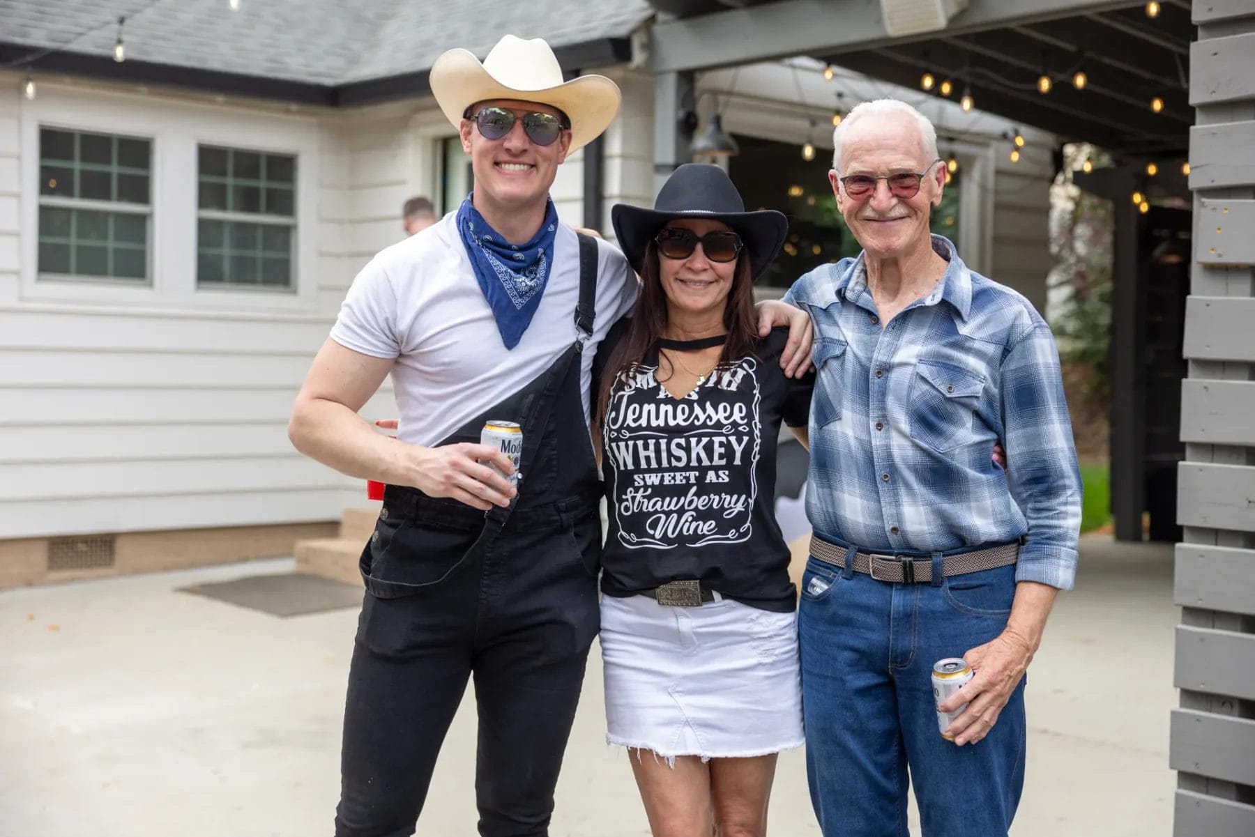In front of a white house adorned with decorative lights, three people smile under the sun. One's in a cowboy hat and overalls, another sports a black hat and T-shirt while sipping a drink, and the third has glasses with a plaid shirt—capturing the spirit perfect for an employee appreciation event in Huntersville, NC. Huntersville event venue