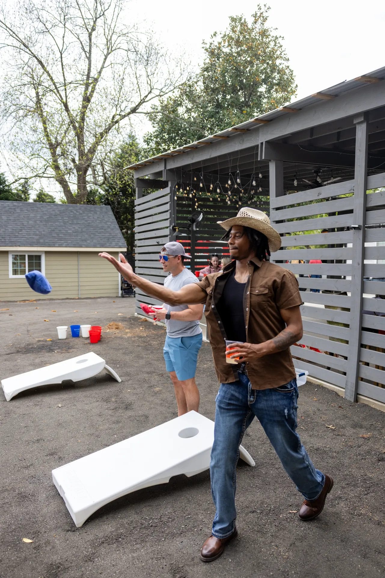 Two men playing cornhole in an outdoor setting. One is casually dressed in a baseball cap and shorts, holding a drink, while the other wears a cowboy hat, jeans, and a brown shirt. Red and blue plastic cups are on the ground. String lights hang above. Huntersville event venue