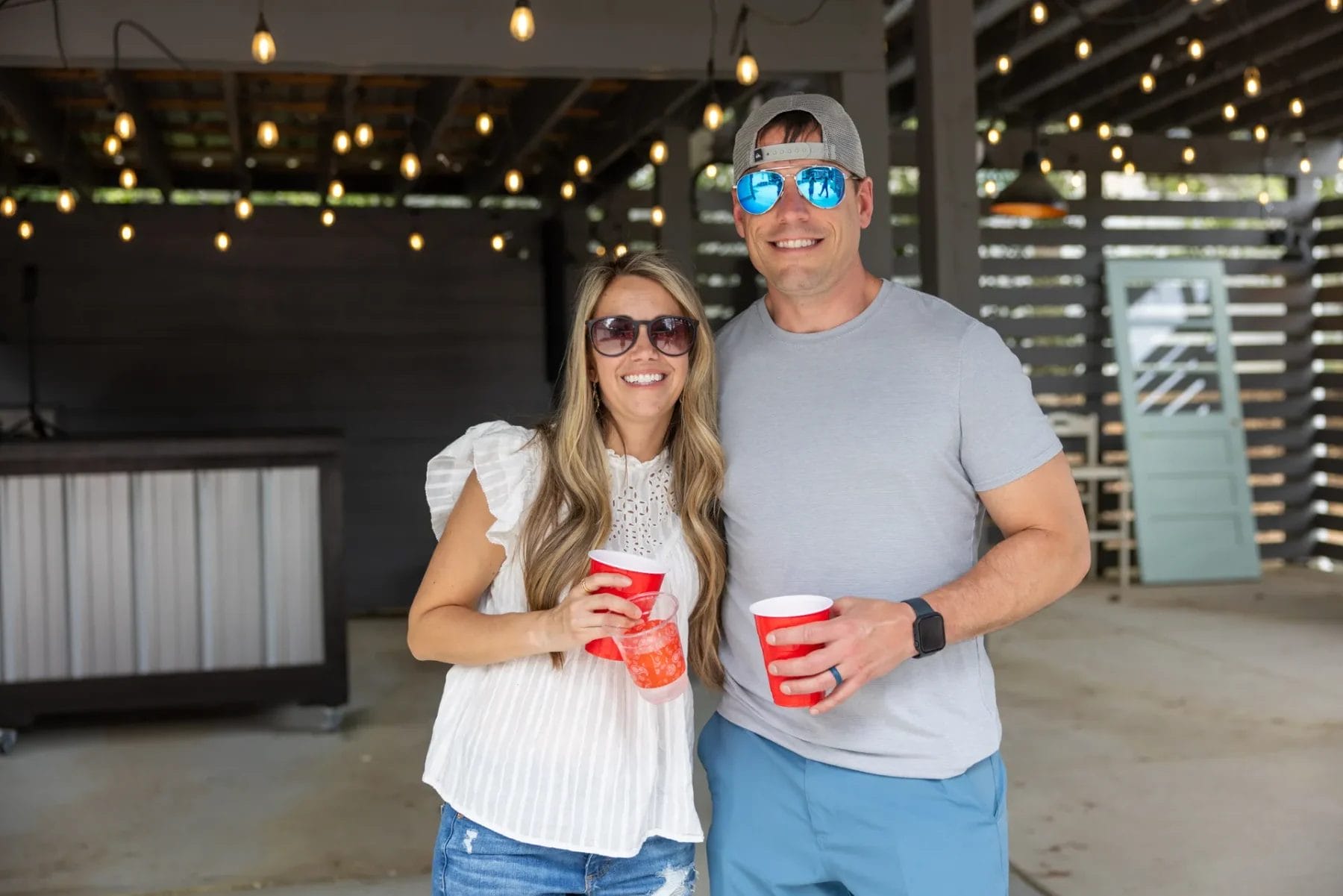 A man and woman stand smiling together outside under string lights. Both wear sunglasses and casual clothing. The woman holds a red cup, and the man holds two. There is a bar and wooden wall in the background. Huntersville event venue
