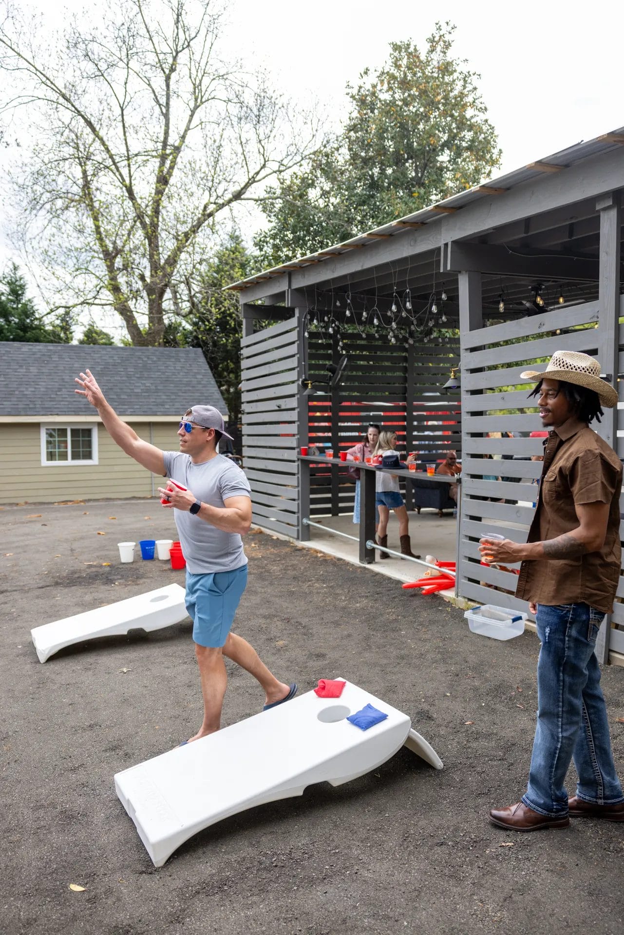 Two men play cornhole outside. One is throwing a beanbag, wearing a gray shirt and cap, while the other stands nearby in a hat and sunglasses. In the background, two people socialize by a covered area with string lights. Huntersville event venue