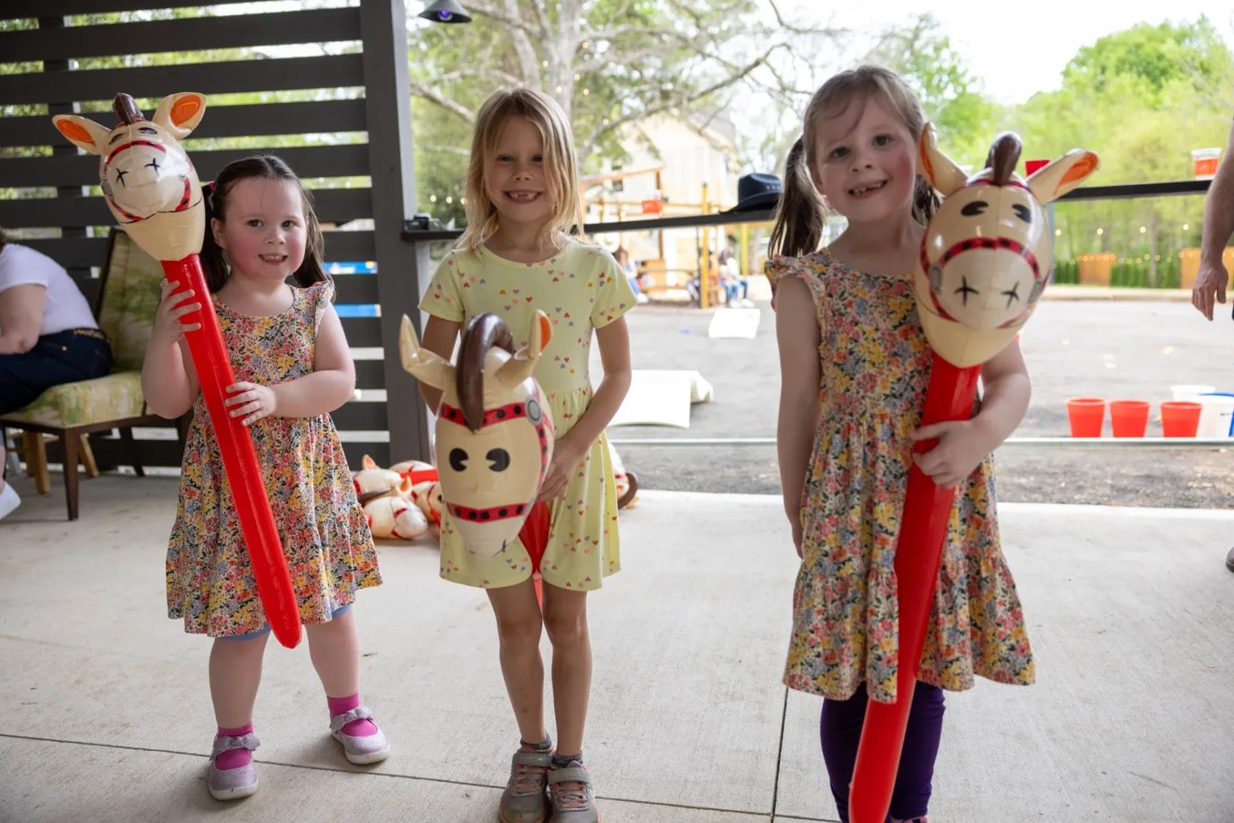 Three children standing indoors, each holding a red hobby horse with a cartoon-like animal head. The children are wearing dresses and smiling. Furniture and outdoor view visible in the background. Huntersville event venue