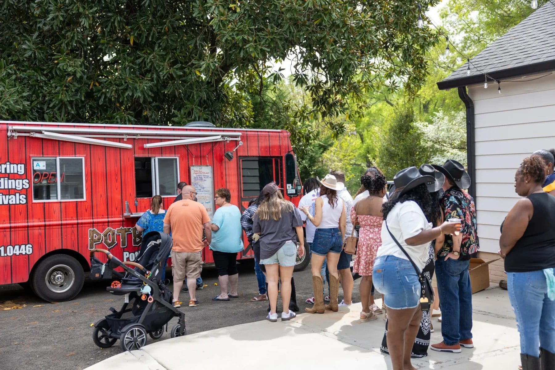 A Heartwarming Office Party to Kick Off 2025 at Manjal Manor 7 A diverse group of people stands in line at a red food truck under large green trees. A stroller is to the side, and the scene includes adults in casual summer clothing, some wearing hats, waiting to order food. Huntersville event venue