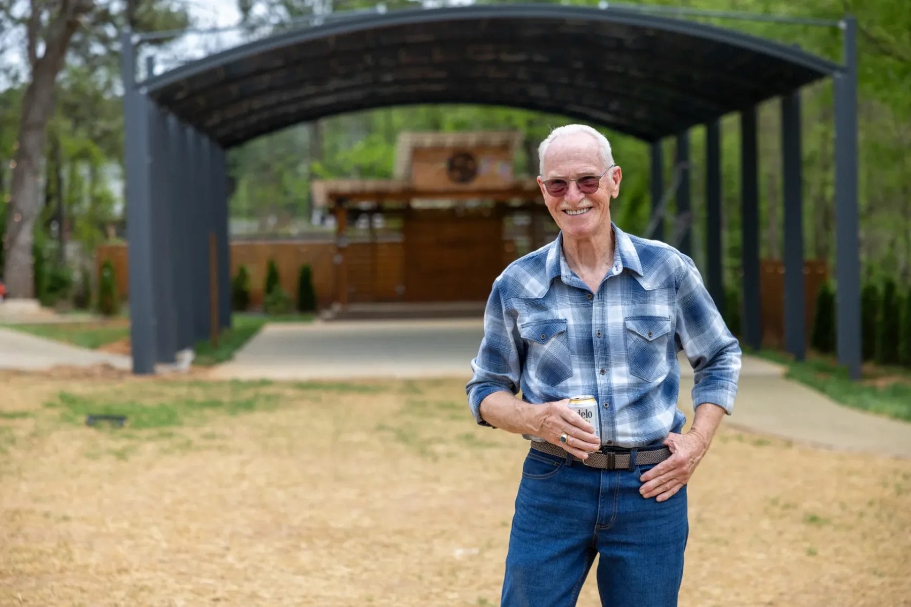 A Heartwarming Office Party to Kick Off 2025 at Manjal Manor 10 An older man in sunglasses stands smiling outdoors, holding a cup. He's wearing a blue plaid shirt and jeans. Behind him is a modern, roofed structure with an open front, set in a grassy area with trees. Huntersville event venue
