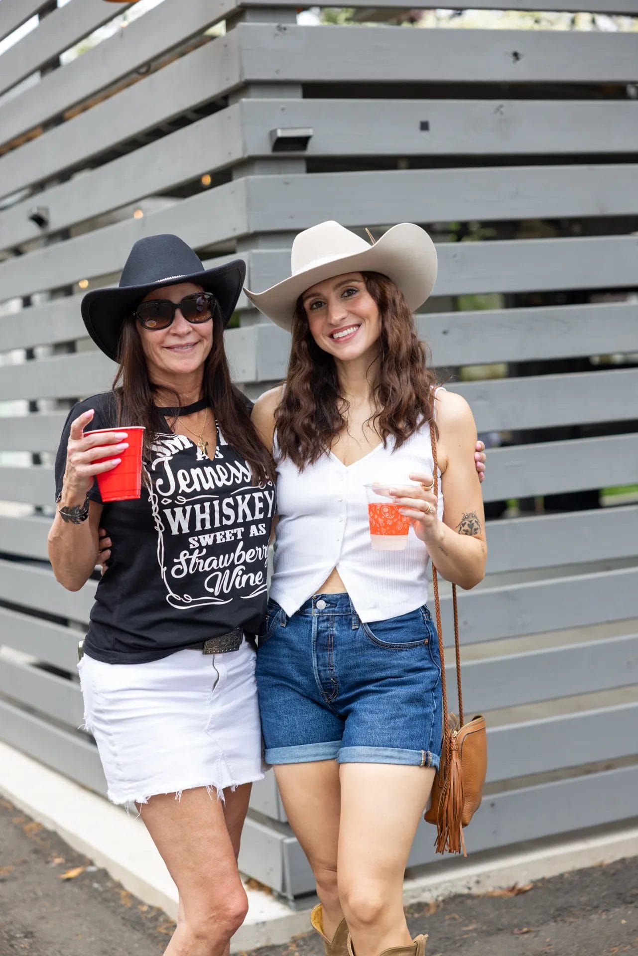 Two women pose together, smiling and wearing cowboy hats. One wears a black t-shirt and white skirt; the other, a white vest and denim shorts. Both hold red cups and stand in front of gray wooden slats. Huntersville event venue