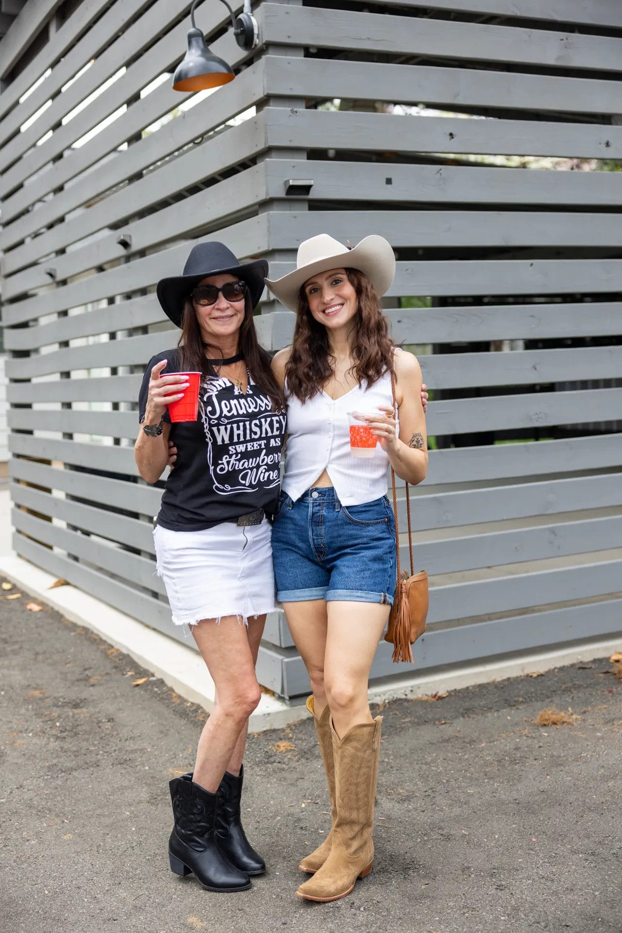 Two women in cowboy hats enjoy drinks at an office party venue in Huntersville. One sports a black hat with a matching shirt and white shorts, while the other opts for a white hat, white top, and denim shorts. Both don cowboy boots as they pose against a slatted wooden wall. Huntersville event venue