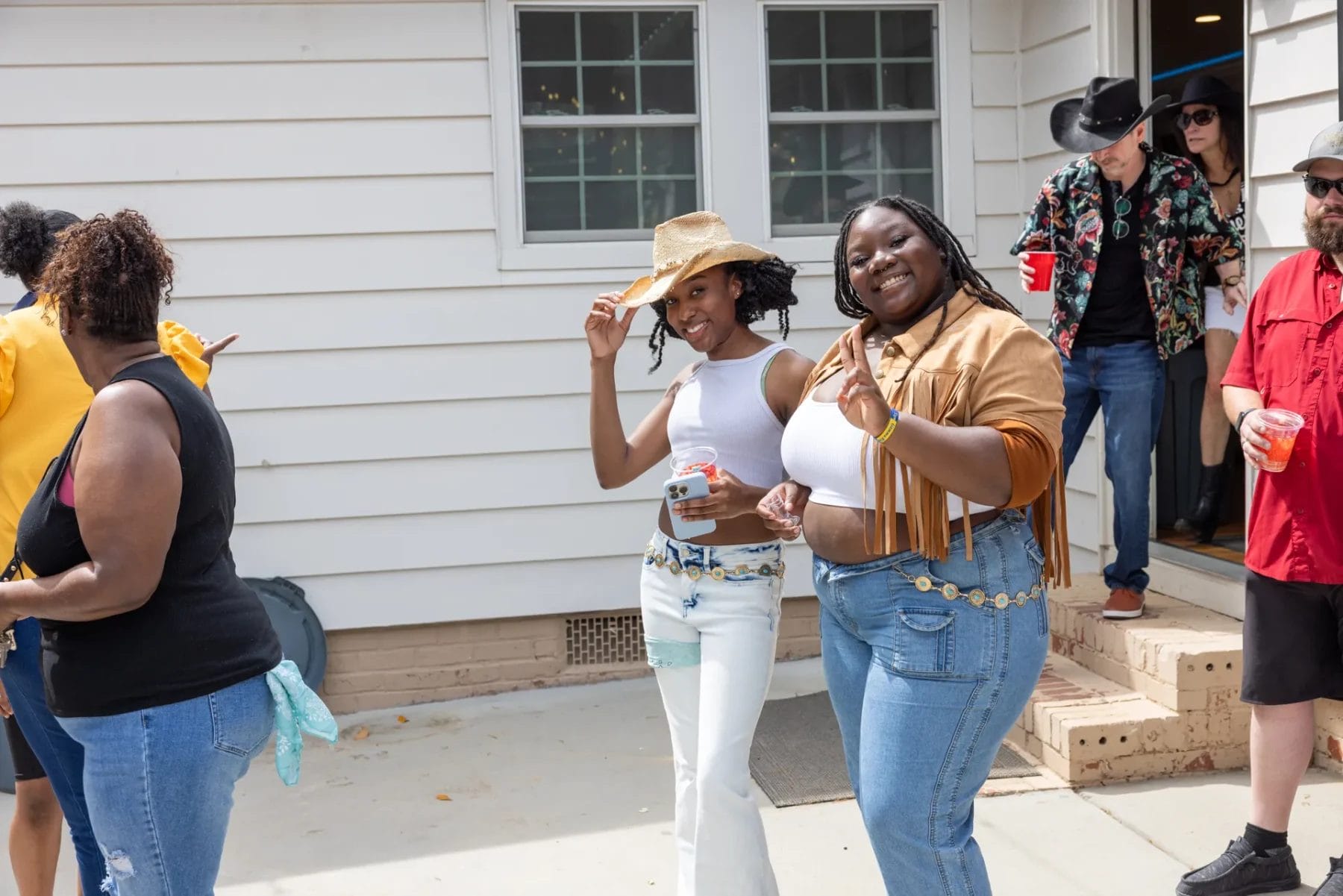 Two people smiling and posing outdoors at a gathering. One wears a straw hat and white top, the other a brown fringe jacket and jeans. Others in the background are mingling and holding drinks. A white building is in the background. Huntersville event venue