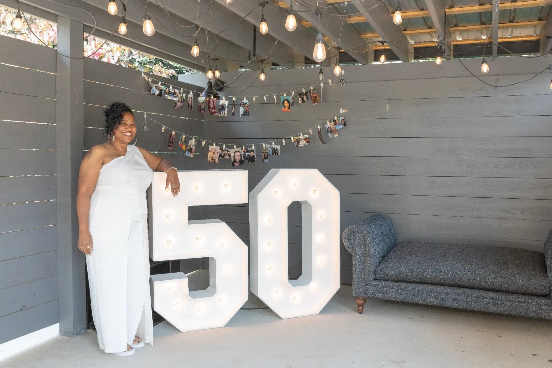 A woman in a white jumpsuit stands next to large, illuminated numbers "50" under hanging lights in a decorated area. Photos are clipped to a string behind her, and a gray couch is on the right. Huntersville event venue