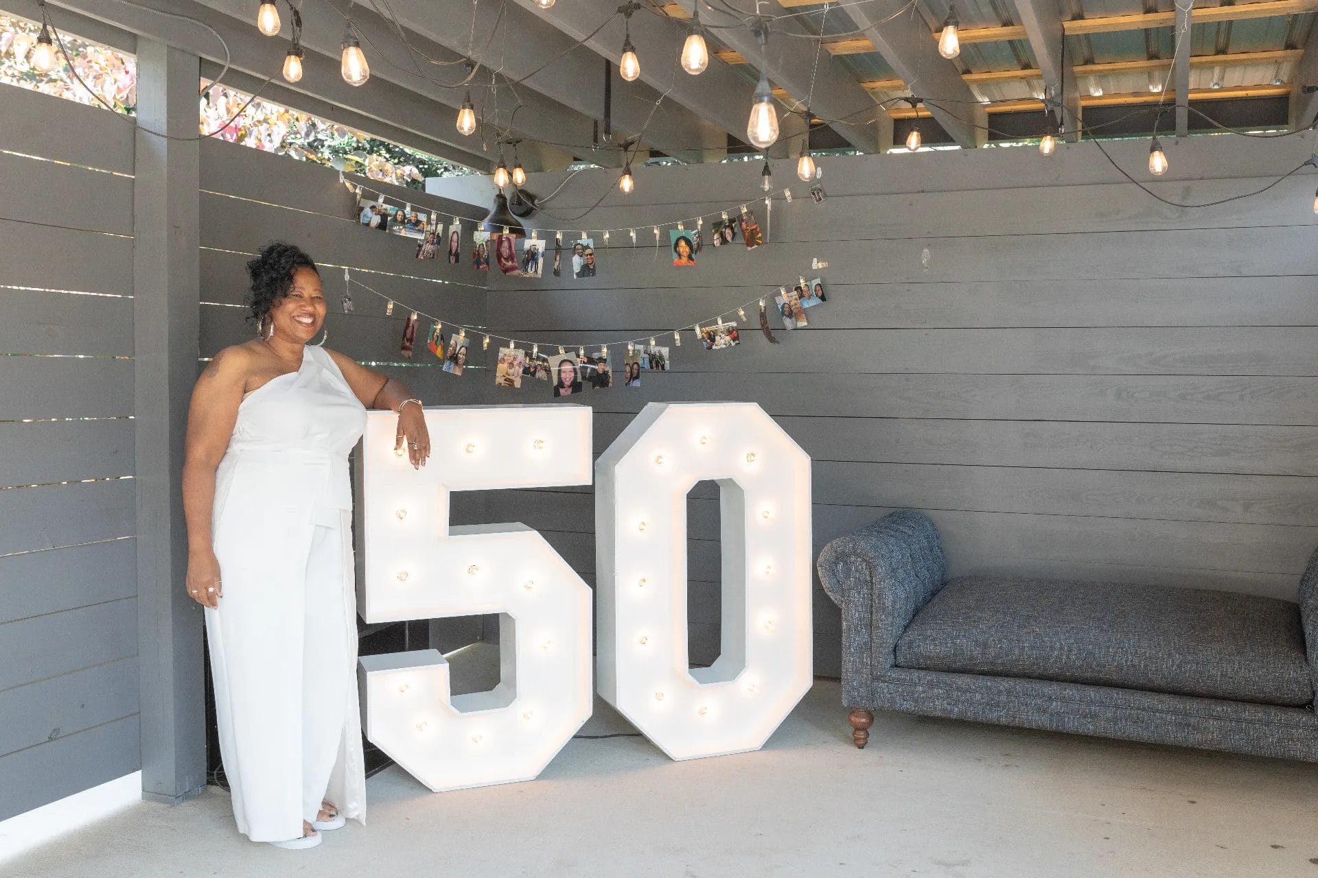 A woman in a white jumpsuit stands next to large, illuminated numbers "50" under hanging lights in a decorated area. Photos are clipped to a string behind her, and a gray couch is on the right. Huntersville event venue
