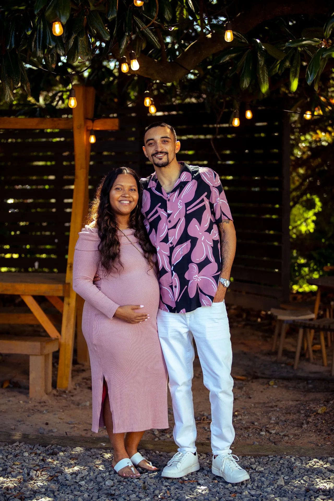 A smiling couple stands outdoors under string lights at a Huntersville baby shower event venue. The pregnant woman, in a pink dress, gently rests her hand on her belly while the man, in a pink patterned shirt and white pants, stands beside her. They're surrounded by lush greenery and wooden structures. Huntersville event venue