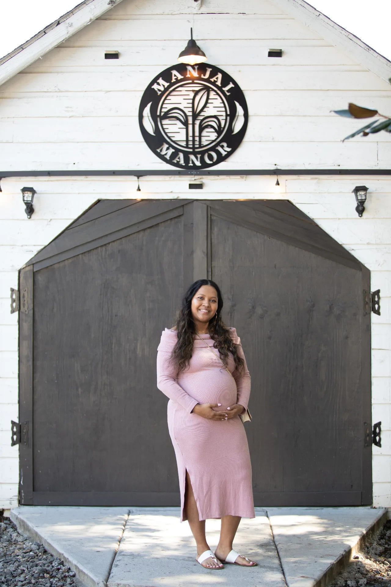 A woman in a pink dress stands with a glowing smile, holding her belly outside the large wooden doors of Huntersville's "Manjal Manor," the perfect baby shower event venue. She is wearing sandals, surrounded by rustic white and dark wood accents that add charm to the joyous occasion. Huntersville event venue