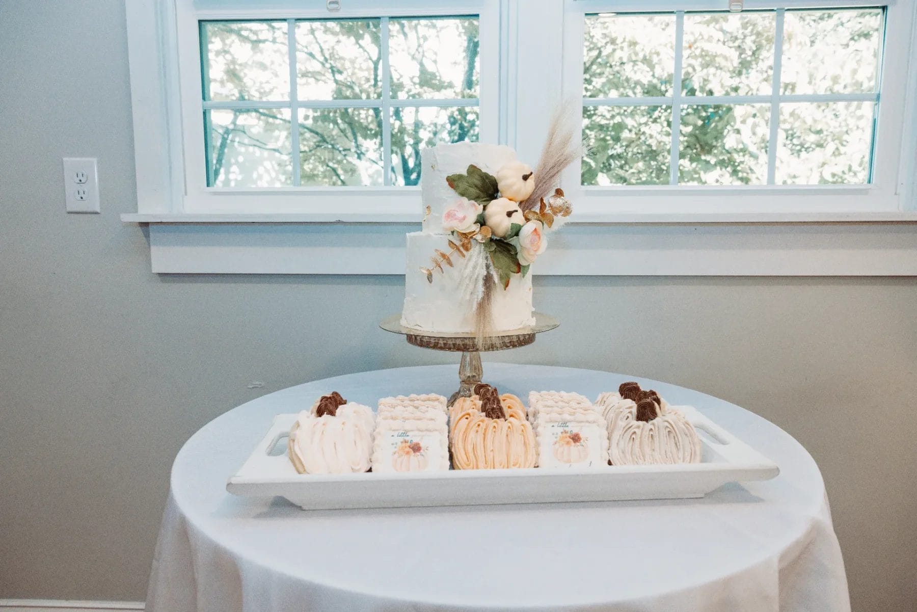 A small, elegant cake with floral decorations sits on a glass stand, perfect for a Huntersville babyshower event. Below it, various bundt cakes are arranged on a white tray. The setup is in front of two windows with sheer curtains, allowing natural light to filter in beautifully. Huntersville event venue
