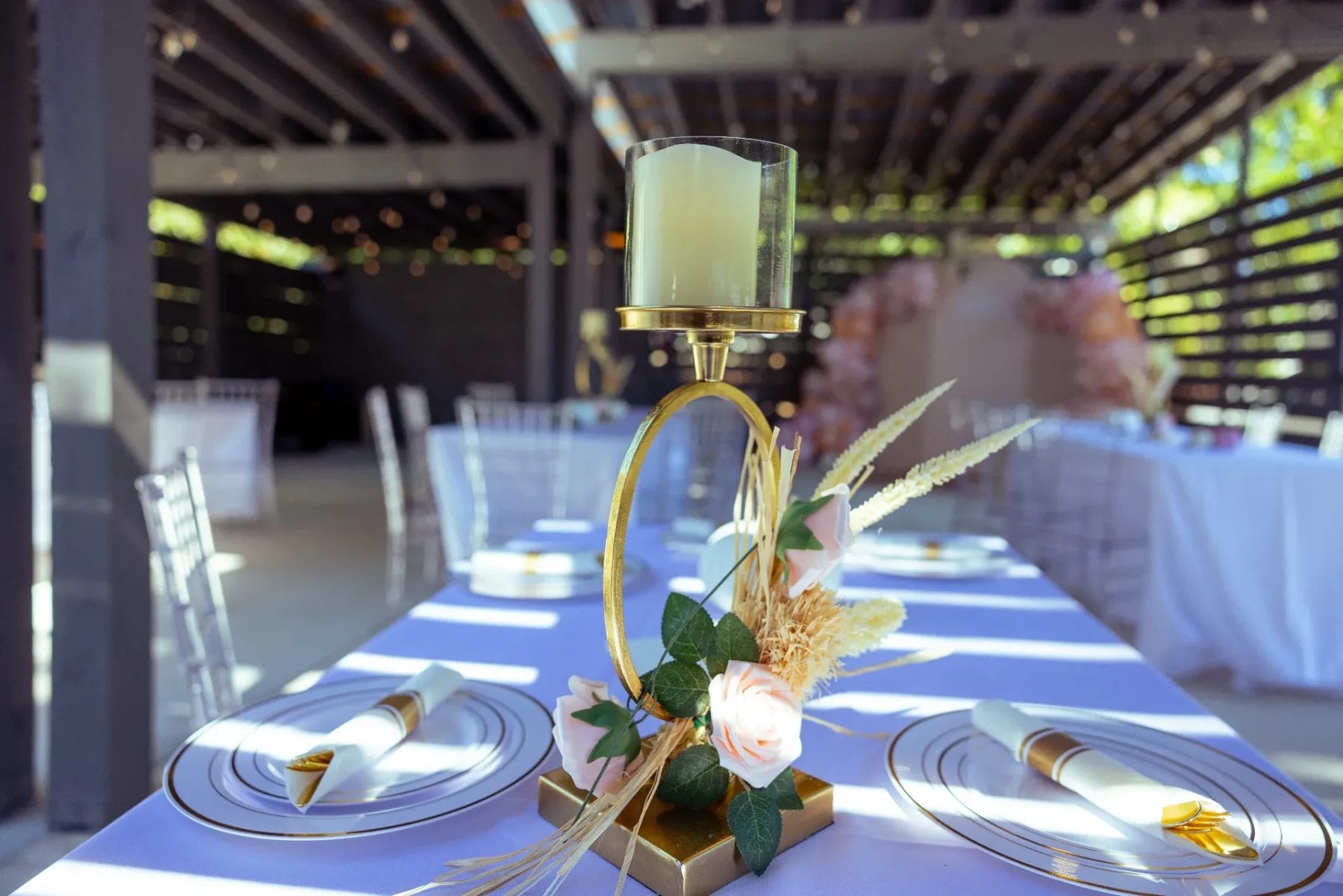 A decorative table setting at a charming Huntersville baby shower event venue features a gold candle holder with a white candle, surrounded by pink flowers and greenery. The table is adorned with white plates and rolled napkins, with sunlight streaming through overhead beams. Huntersville event venue