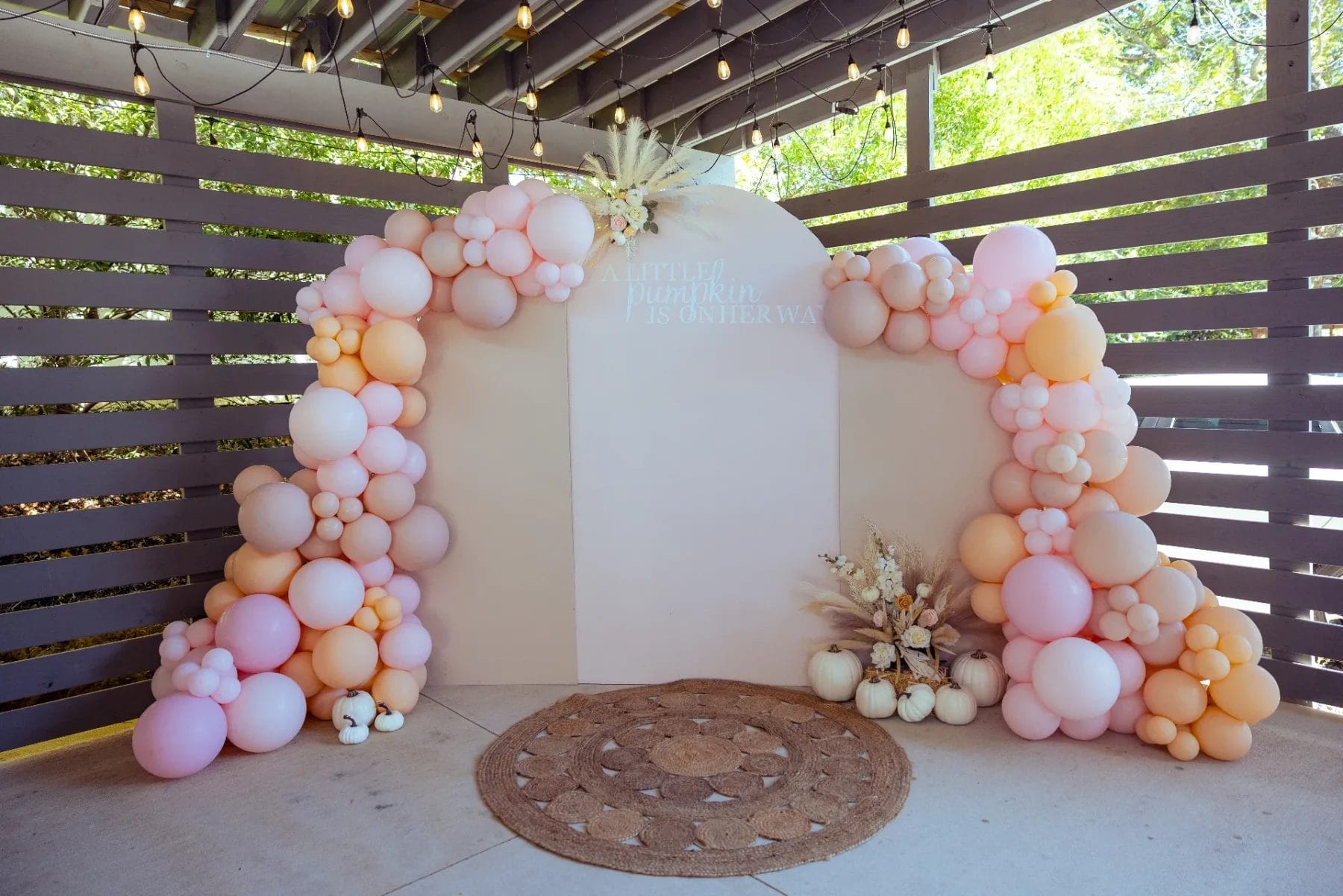 A decorative archway with pastel balloons in shades of pink, peach, and cream, adorned with small pumpkins and dried flowers. A round woven rug lies in front under a wooden pergola with string lights at the charming Huntersville baby shower event venue. Text reads "A little pumpkin is on the way. Huntersville event venue