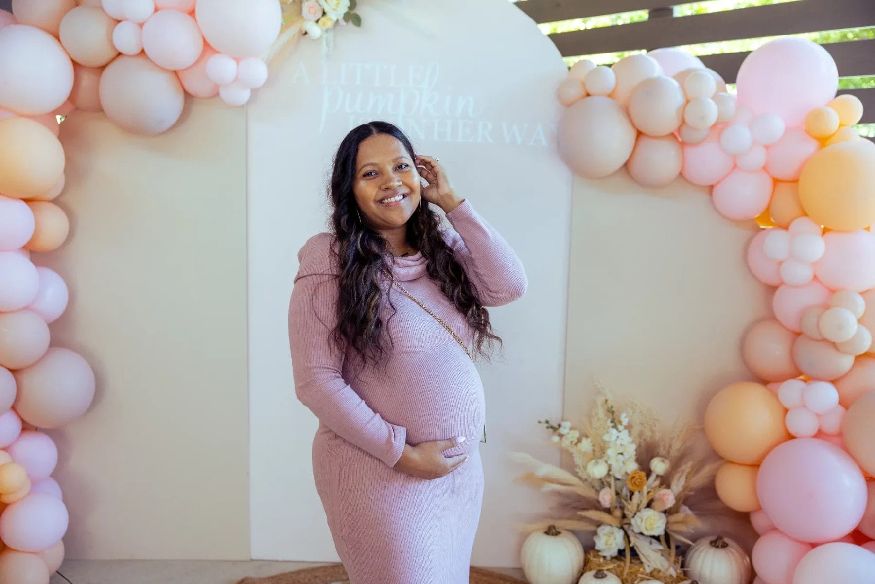 A pregnant woman in a pink dress stands smiling at her baby shower at a charming Huntersville venue. She is surrounded by pastel-colored balloons, with a backdrop that says, "A Little Pumpkin is on Her Way." Dried flowers and pumpkins elegantly decorate the scene. Huntersville event venue