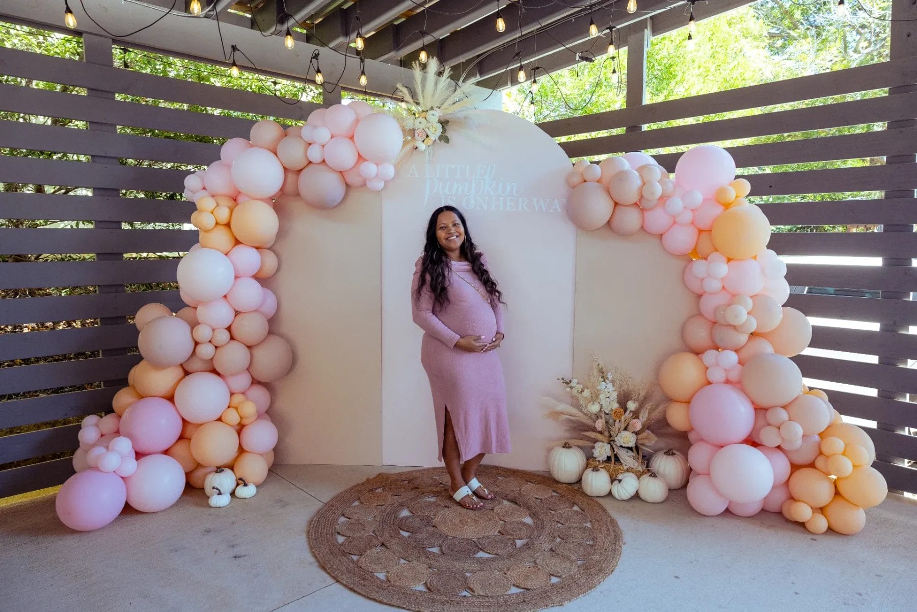 A pregnant woman in a pink dress stands smiling at a Huntersville babyshower event venue, framed by a festive backdrop with a balloon arch. The setup features pink, peach, and cream balloons, a circular straw rug, and charming small white pumpkins. Huntersville event venue