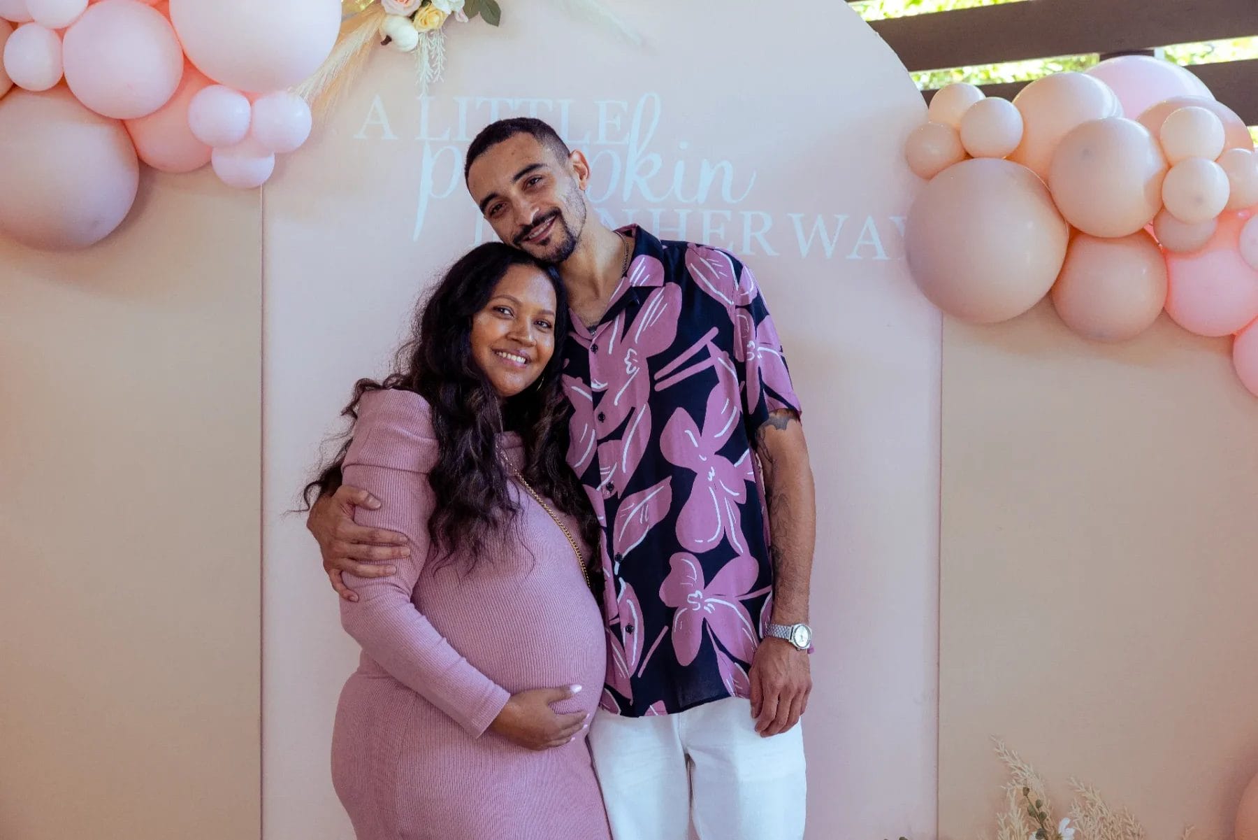 A pregnant woman in a pink dress stands next to a man in a floral shirt, both smiling at the Huntersville Babyshower Event Venue. They are in front of a peach-colored backdrop with pink balloons and the words "A little pumpkin is on her way" displayed behind them. Huntersville event venue