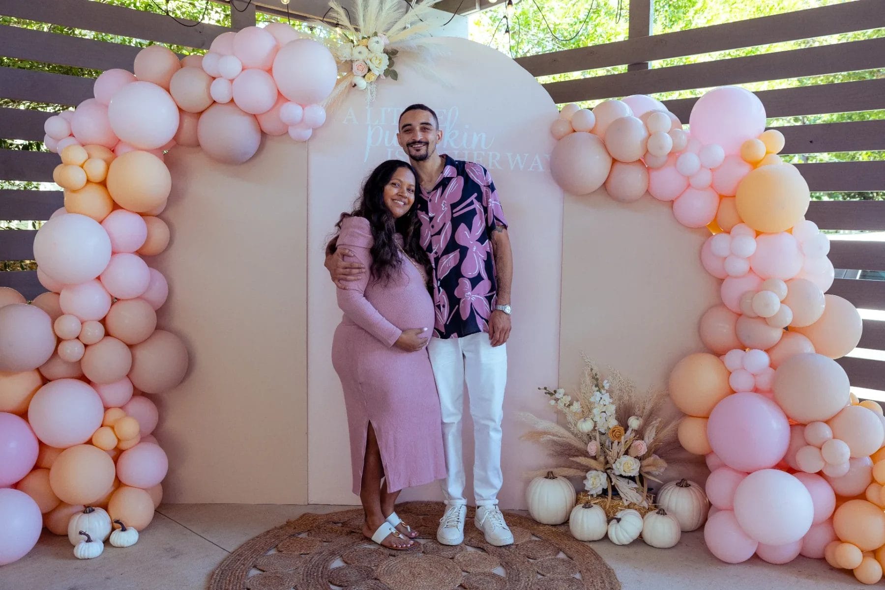 A couple stands in front of pink and peach balloon arches at a Huntersville babyshower event venue, with dried floral arrangements and white pumpkins at their feet. The woman cradles her baby bump, both smiling at the camera in this festive setting. Huntersville event venue