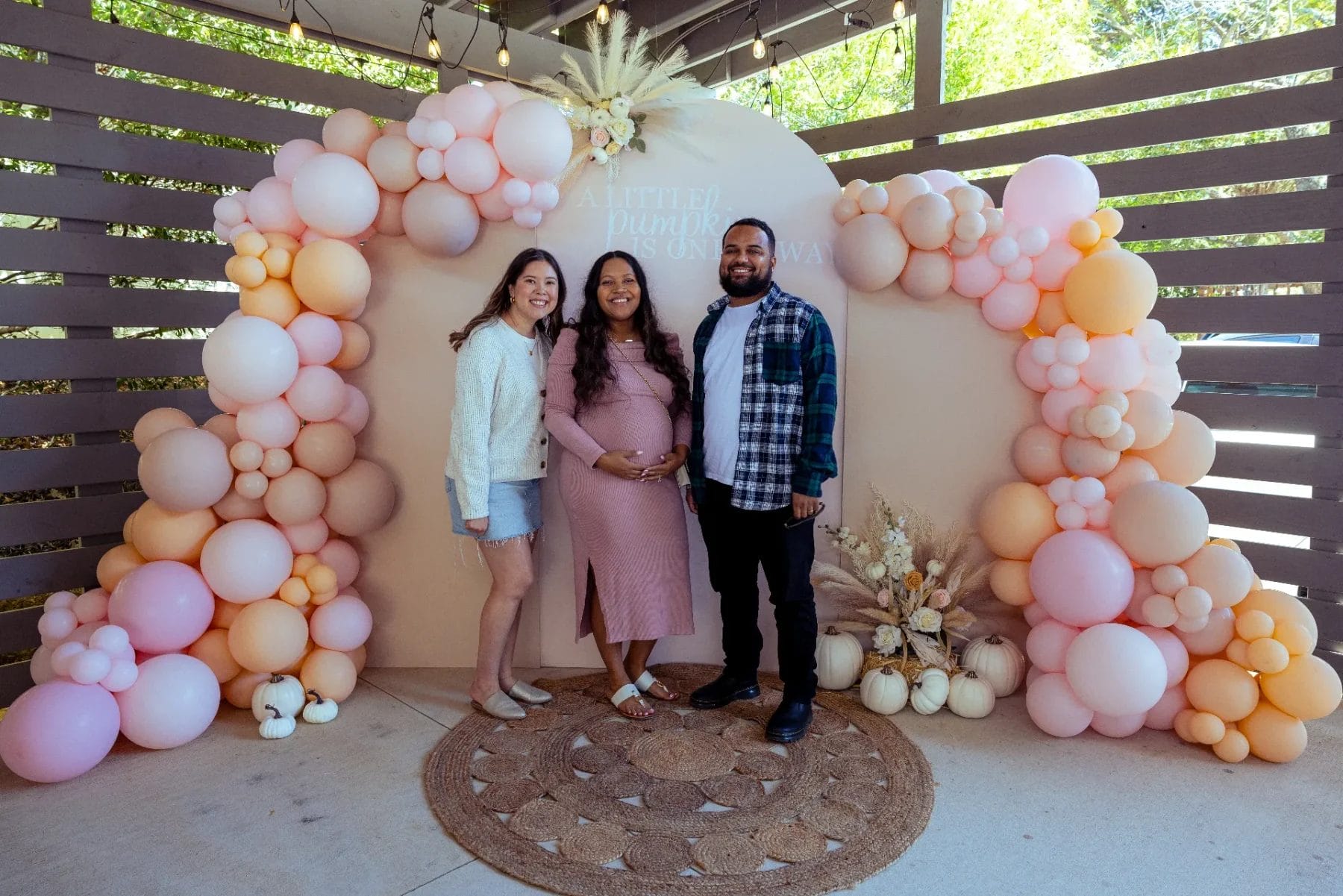 Three people stand smiling at a baby shower in Huntersville. The expectant person in the center wears a pink dress and holds their belly, surrounded by pastel balloon decorations and small pumpkins on the ground, making the venue perfect for celebrating new beginnings. Huntersville event venue