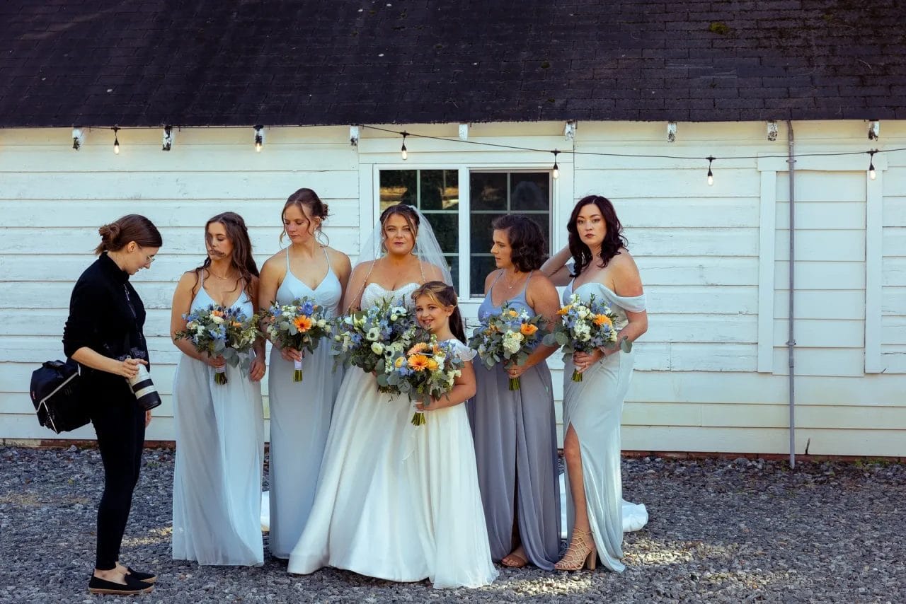 A bride in a white dress stands with five bridesmaids in light gray dresses holding bouquets. They are posing for a photographer outside, in front of a rustic white building with string lights. Huntersville event venue