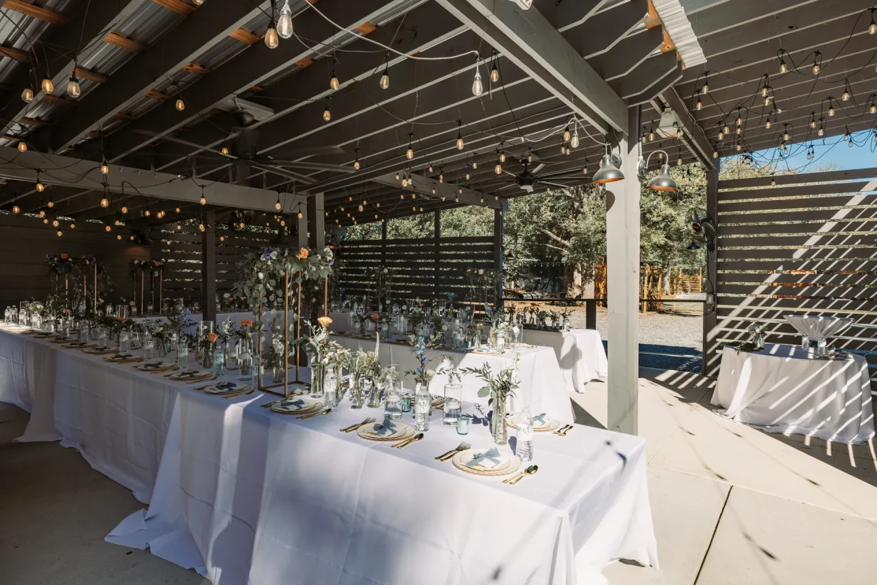 A beautifully decorated outdoor wedding reception area with long tables covered in white tablecloths. Each table is adorned with elegant floral arrangements and dining settings. String lights hang from the ceiling, creating a warm, inviting atmosphere. Huntersville event venue