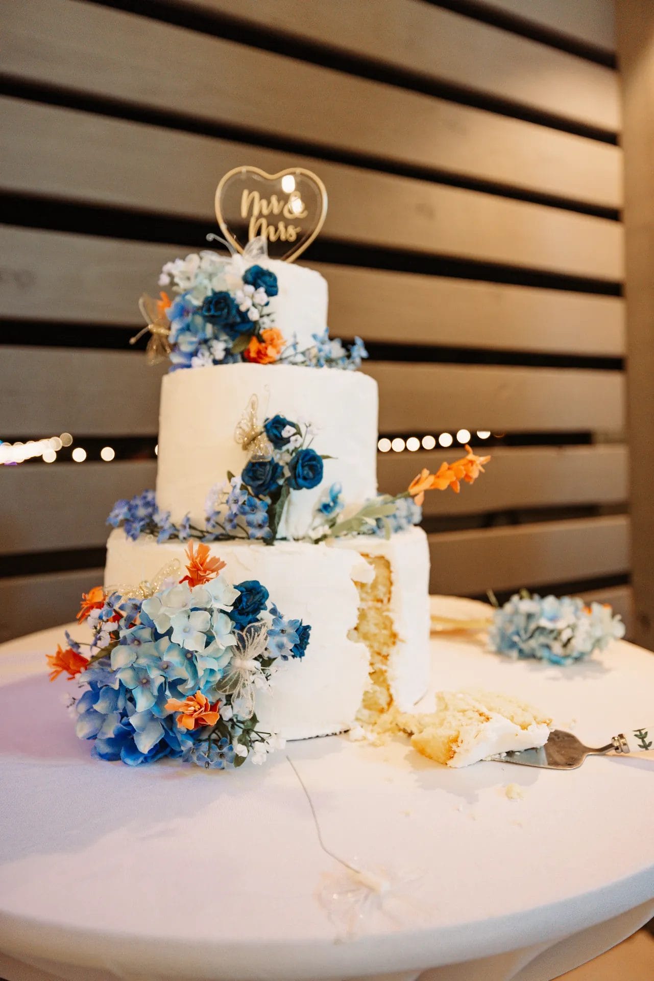 A three-tiered white wedding cake decorated with blue and orange flowers sits on a table. A slice has been removed, and a cake server is lying beside it. A "Mr. & Mrs." topper is on the cake. Huntersville event venue