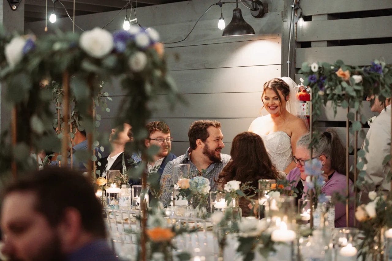 A bride in a white gown smiles and interacts with seated guests at a decorated wedding reception. The setting includes floral arrangements, candles, and soft lighting, creating a warm and festive atmosphere. Huntersville event venue