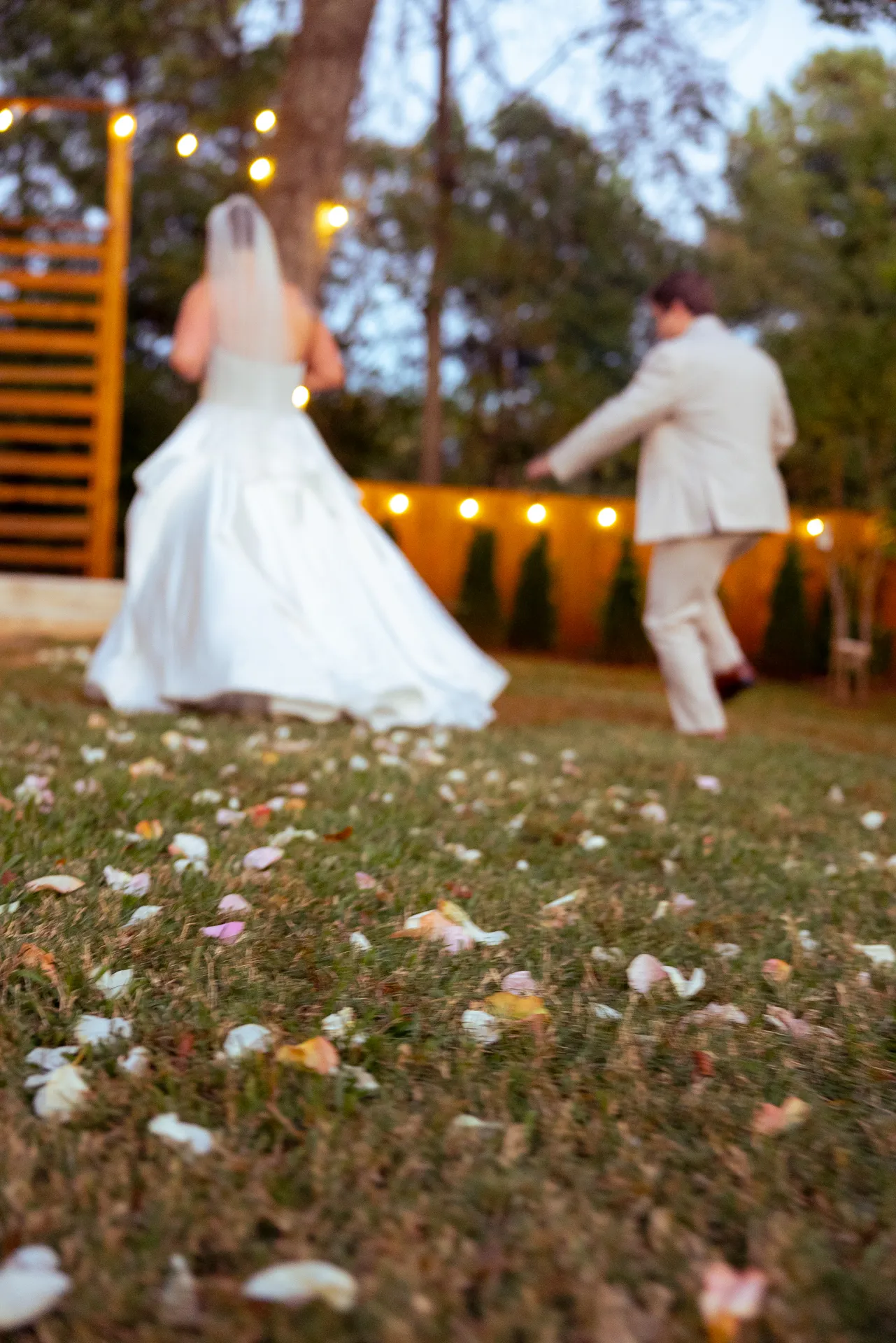 A bride and groom joyfully walking over a grass field adorned with scattered flower petals. The bride wears a white gown and veil, and the groom is in a light suit. String lights and trees form the background, creating a festive atmosphere. Huntersville event venue